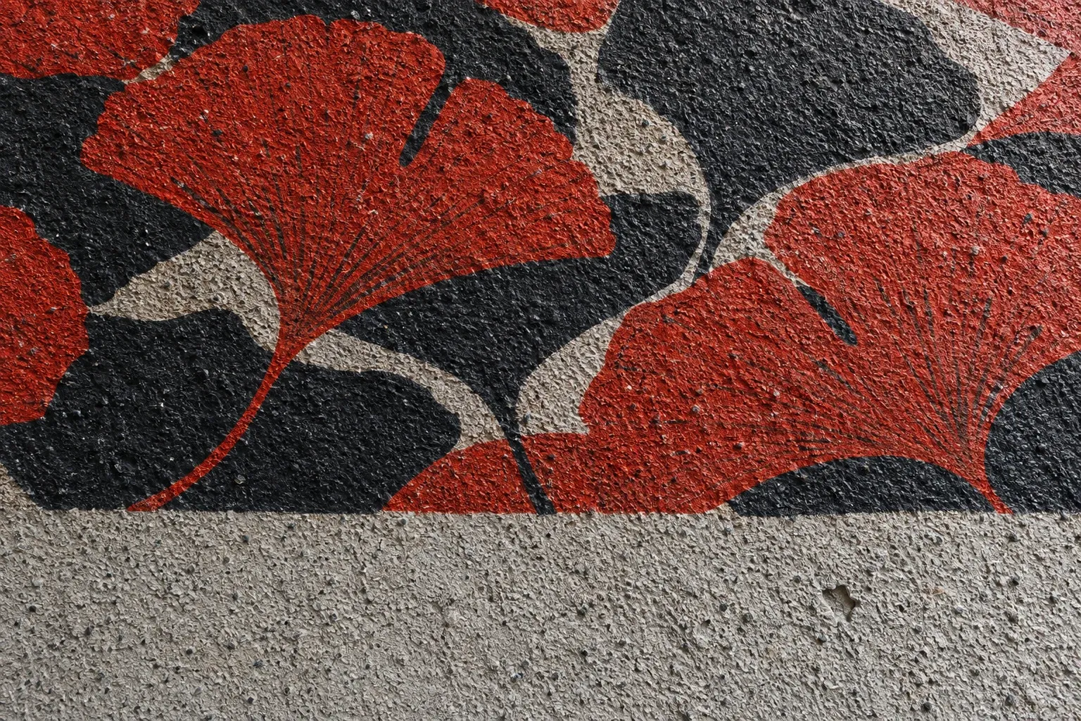 Crépi à gros grain en lumière rasante, feuilles de ginkgo gris anthracite et vermillon imprimées dans le relief, bas du mur laissé brut